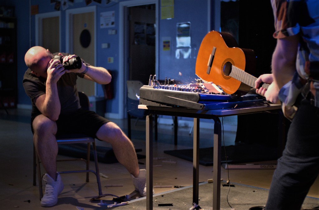 Photographer takes a photo of man smashing an acoustic guitar over a mixing desk.